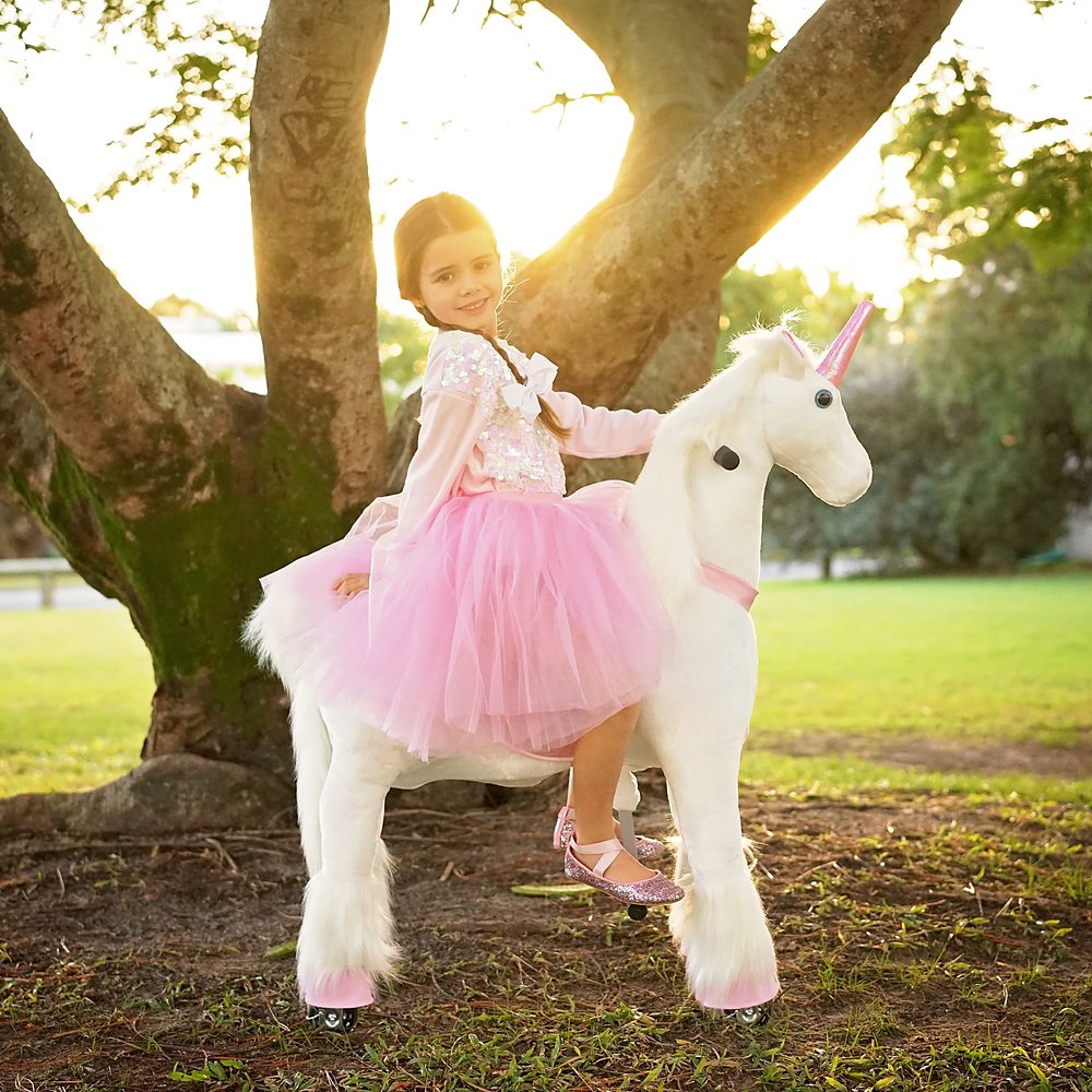 Little Riders pink and white unicorn ride-on toy - girl riding her pink and white unicorn under the tree