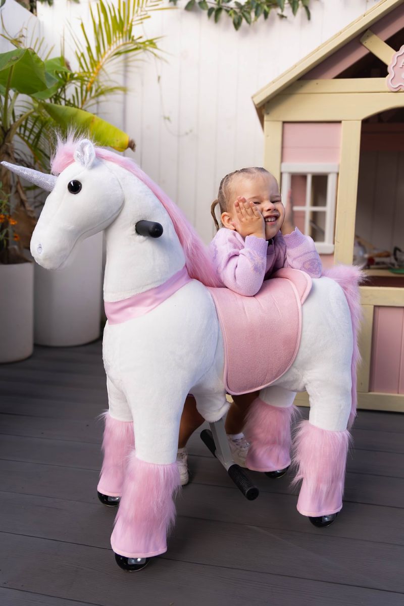 Little Riders pink unicorn ride-on toy - toddler smiling over her pink unicorn by the cubby house